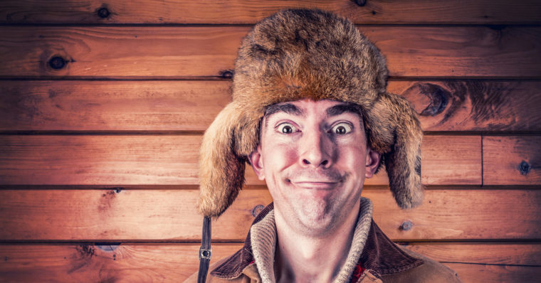 Photo of a man posing in front of a pine-wood wall, wearing a furry hat, and making a silly face.