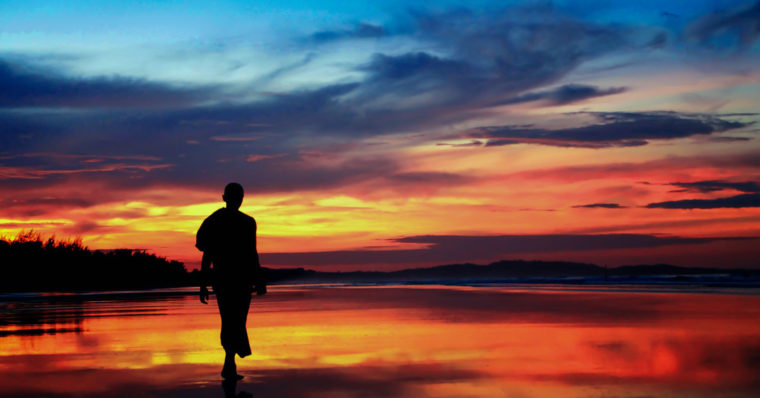 A photo of a man walking peacefully on the beech at sundown.