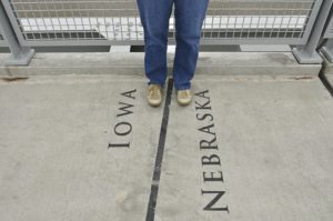 A person's feet, straddling a state borderline between Iowa and Nebraska, to play on the idea of being in two places at once.