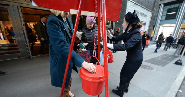 Photo of a Salvation Army bell ringer next to her red kettle, with a donor chipping in. 